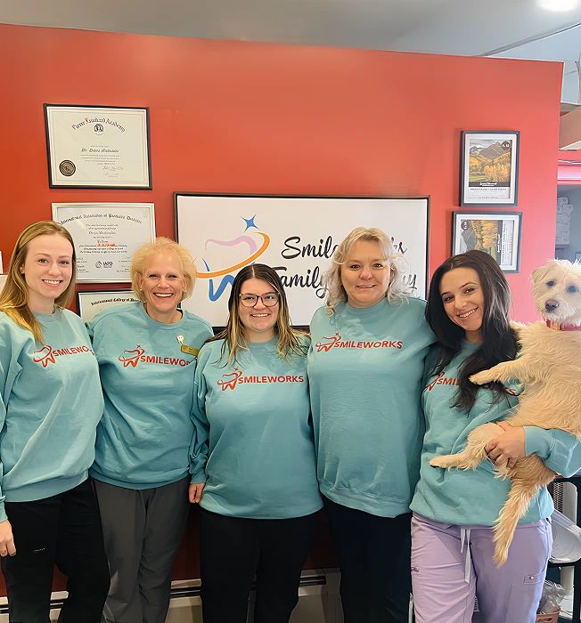 Five Smileworks team members in matching sweatshirts, one holding a small dog, in front of the clinic’s logo wall